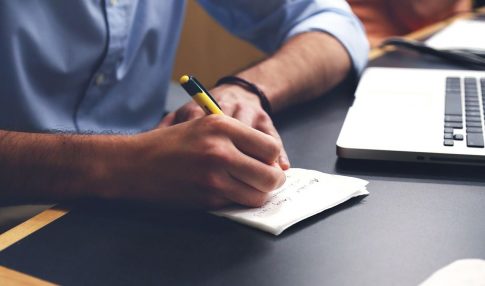 A person writing on a piece of paper next to a laptop.